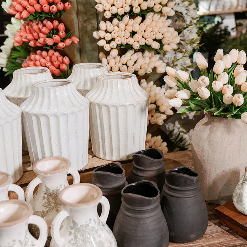 Collection of ceramic vases and pots on a wooden surface with floral arrangements in the background.