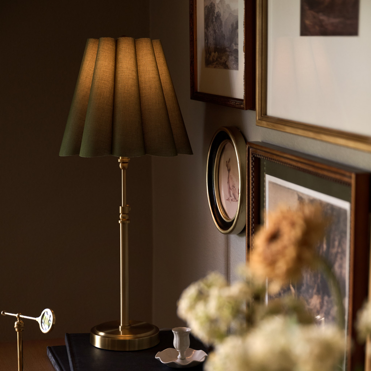 A table lamp with a pleated olive-green fabric shade and a polished brass base stands on a dark tabletop in a styled interior. Framed wall art, a round clock, and blurred flowers are visible in the background and foreground, creating a warm, cozy atmosphere.