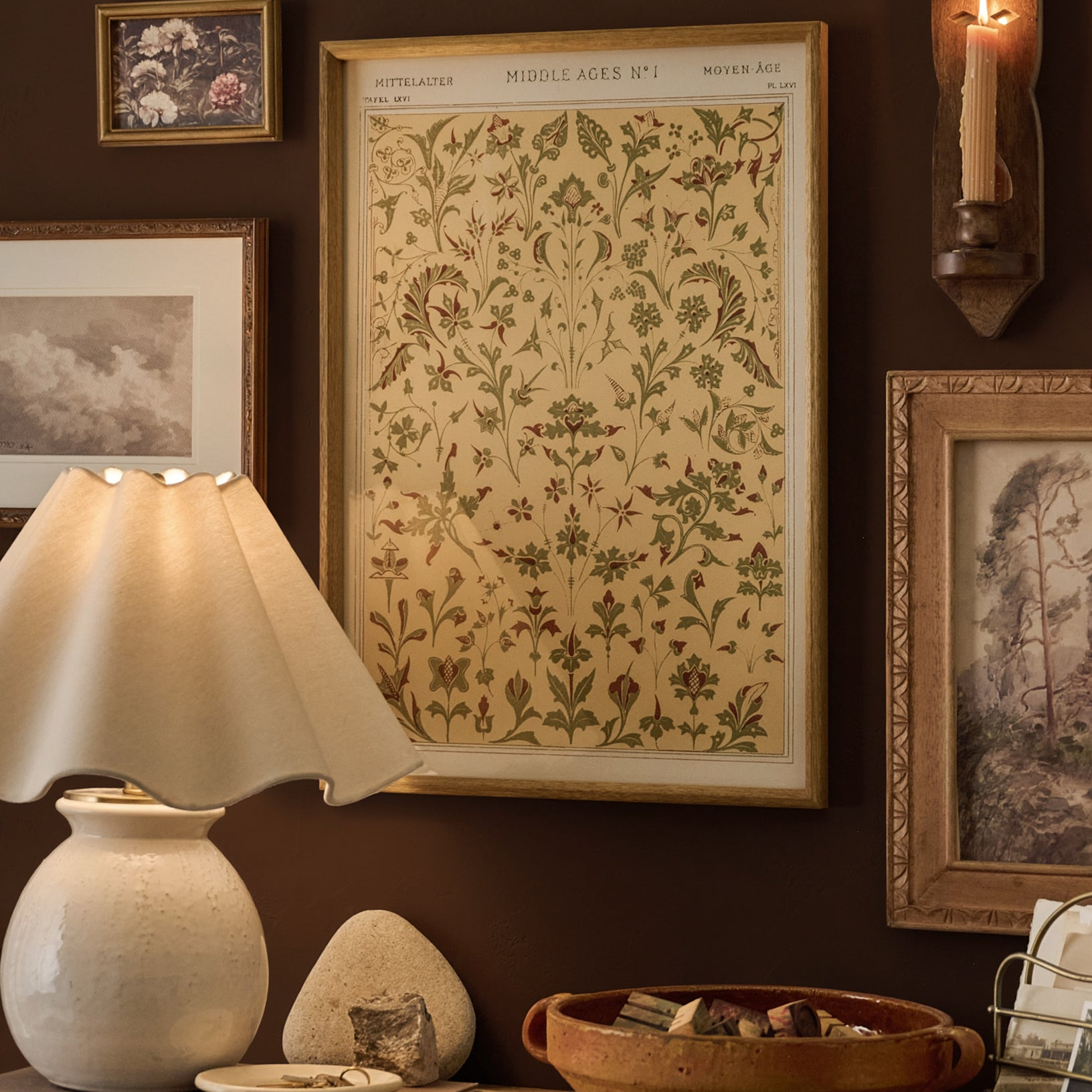 A framed vintage botanical print titled 'Middle Ages No. 1' hangs on a dark brown wall in a styled room setting with a lit table lamp, ceramic objects, and other framed artwork.