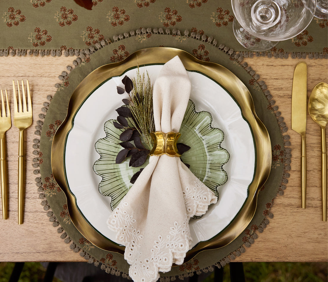 An overhead styled table setting featuring a scalloped gold charger plate with a green and white patterned dinner plate, a cream eyelet napkin tied with a gold napkin ring, and gold flatware on a wooden table.