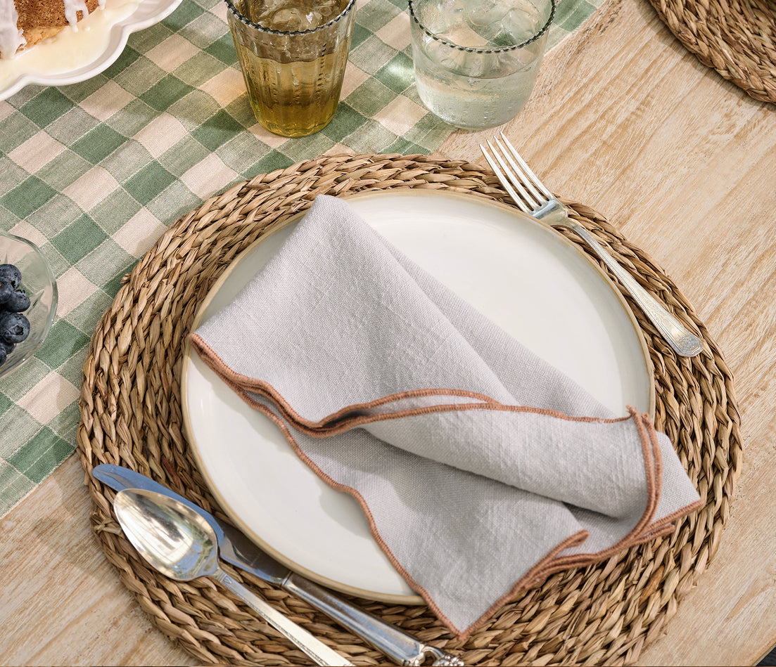 A styled table setting featuring a round woven straw placemat, a white ceramic plate, a folded light gray linen napkin with tan edging, silver cutlery, and a green-and-white checkered tablecloth.