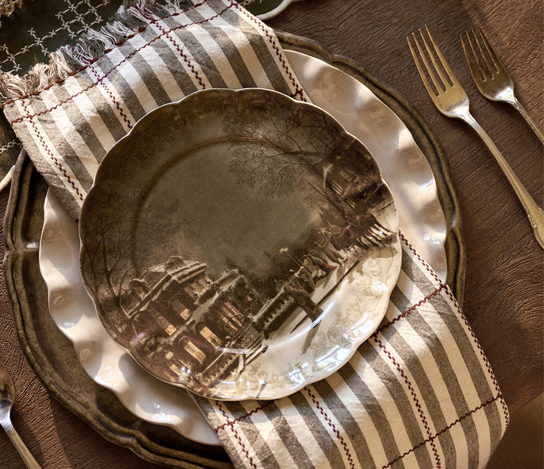 A styled table setting featuring a scalloped ceramic plate with a sepia-toned architectural landscape design, layered over a white charger plate and a striped beige and brown napkin on a dark wood table, with silver forks nearby.