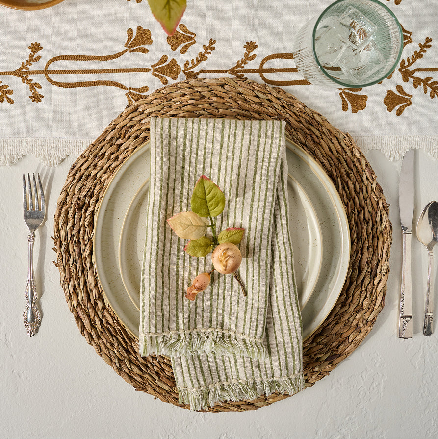 A styled table setting featuring a woven round placemat, layered with cream plates, a striped linen napkin with fringe, and a small leaf sprig garnish, on a white embroidered tablecloth.