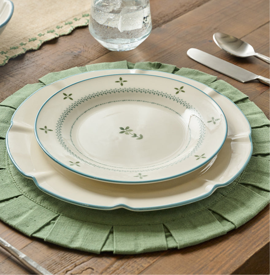 A set of three ceramic plates with a cream glaze, decorated with green floral and dotted border patterns, styled on a green fringed placemat on a wooden table.