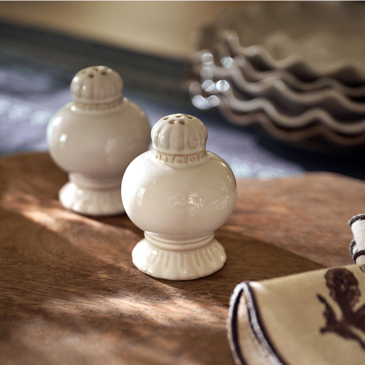 Two small glossy ceramic salt and pepper shakers in a warm off-white or cream glaze, placed on a wooden surface with a softly blurred dining setting in the background.