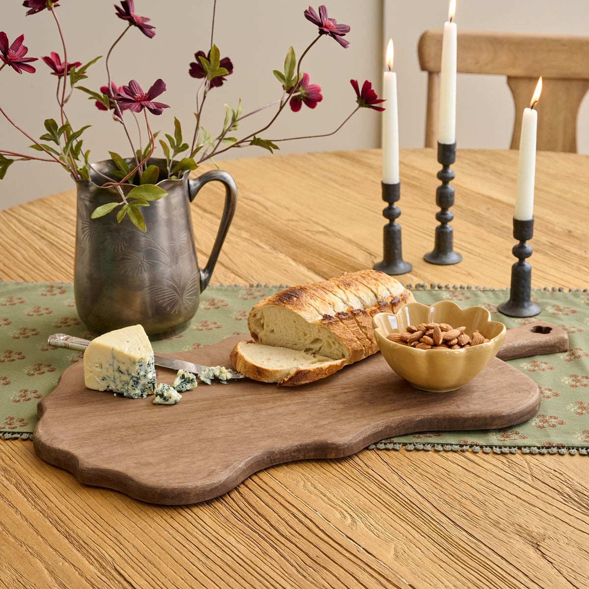 An irregular-shaped wooden serving board styled on a wooden dining table with a loaf of bread, blue cheese, a small bowl of nuts, and a metal pitcher with flowers, alongside three lit candles in black candlesticks.