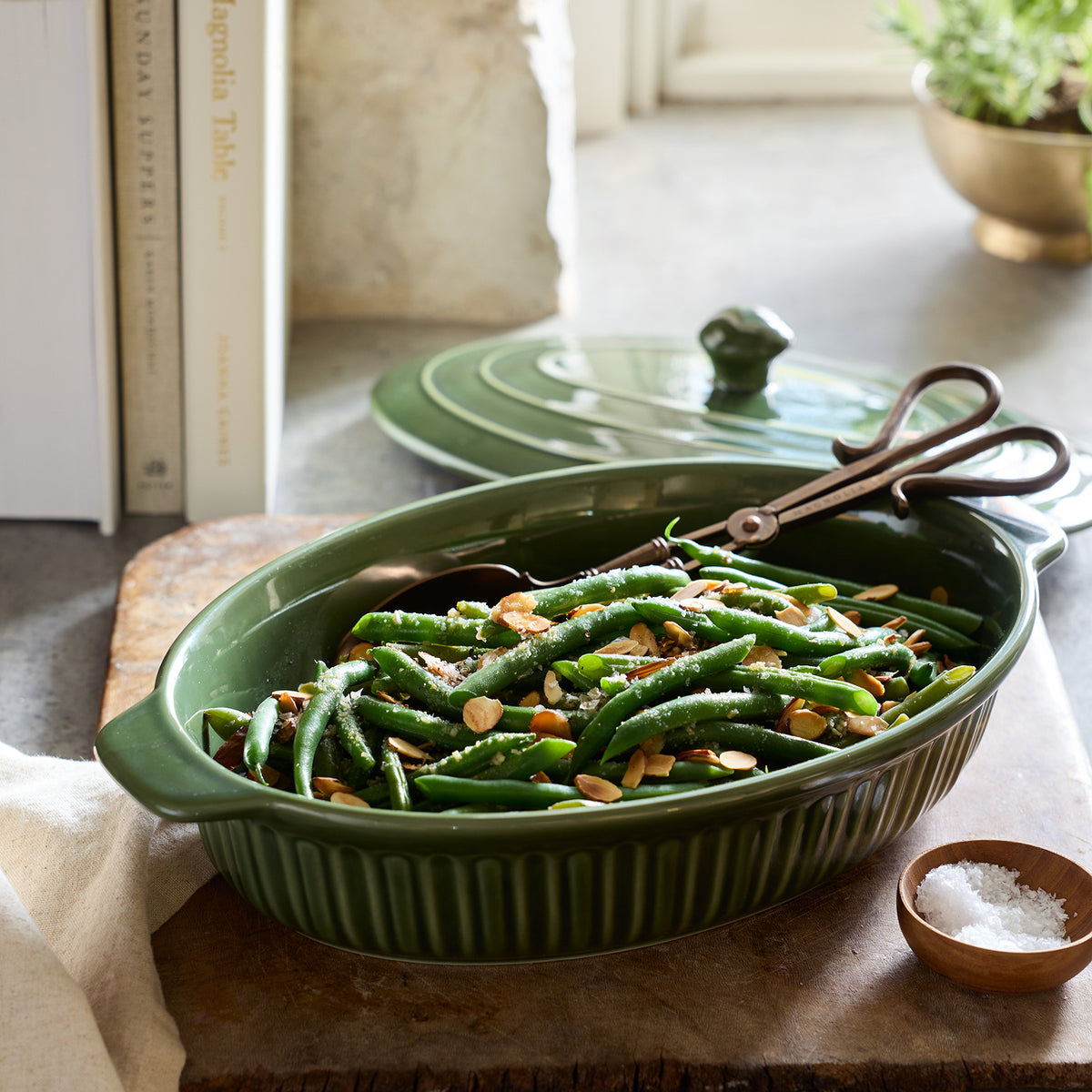 A ribbed oval green ceramic casserole dish filled with cooked green beans and sliced almonds, shown on a wooden board with the matching lid and serving tongs behind it. A small bowl of salt sits nearby in a kitchen-like setting.