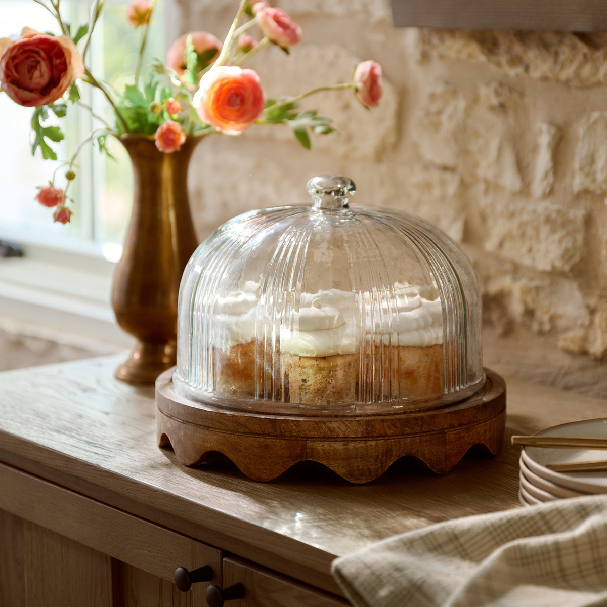 A clear ribbed glass dome cake cover with a small silver knob, resting on a scalloped wooden base, covering cupcakes on a wooden table in a rustic kitchen setting.