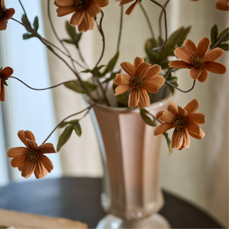 Artificial orange daisy flowers with green leaves arranged in a metallic rose gold vase on a dark table against a soft beige curtain background.