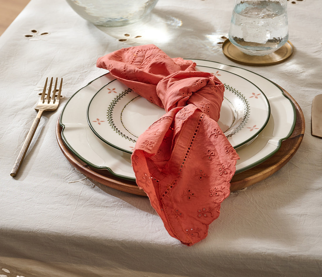A coral pink embroidered cotton napkin tied in a knot, displayed on a place setting with white patterned plates, a gold charger plate, silver cutlery, and a white tablecloth.