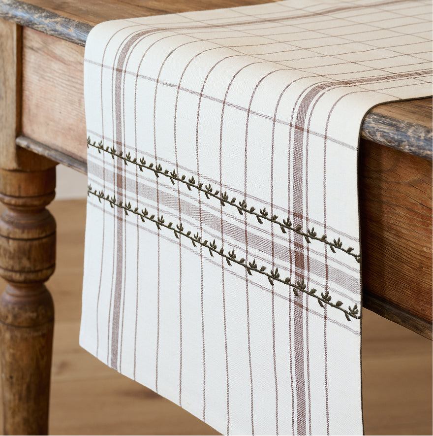 A cream-colored table runner with brown stripes and a leafy vine border, draped over a rustic wooden table.