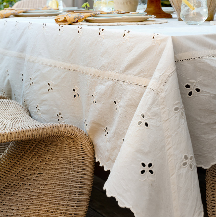 An off-white cotton eyelet tablecloth with embroidered floral and geometric patterns draped over a dining table, styled with plates and glasses, with a wicker chair in the foreground.