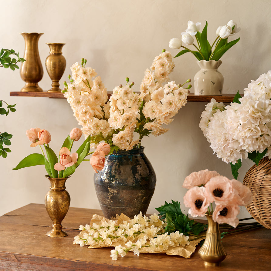 A styled floral arrangement scene on a wooden table with multiple artificial flowers in vases. The central and most prominent item is a bouquet of cream-colored artificial flowers in a dark blue-gray vase, surrounded by peach tulips in a gold vase, white hydrangeas in a wicker basket, and additional flowers on a shelf above.