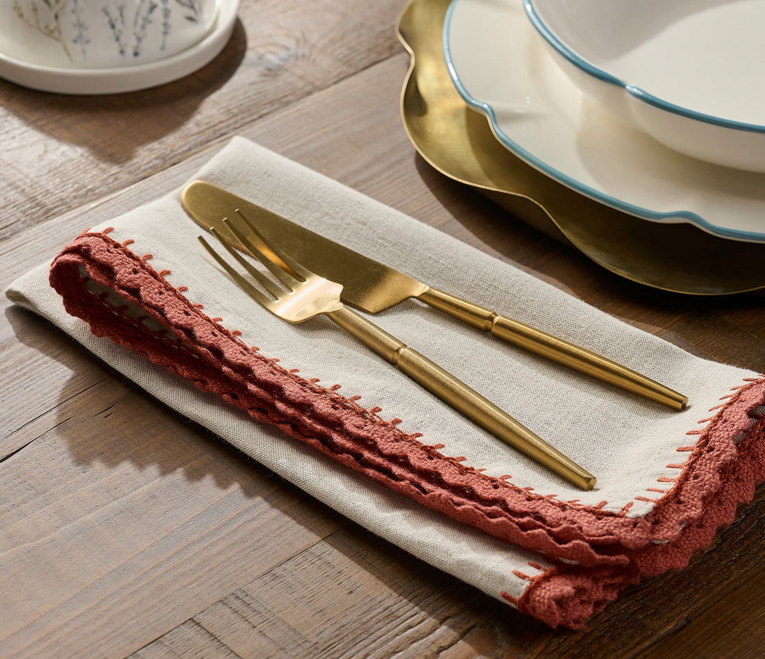 Gold flatware with a matte finish, including a knife and fork, arranged on a folded cream linen napkin with a rust-red embroidered border, set on a rustic wooden table next to a gold and white ceramic plate.