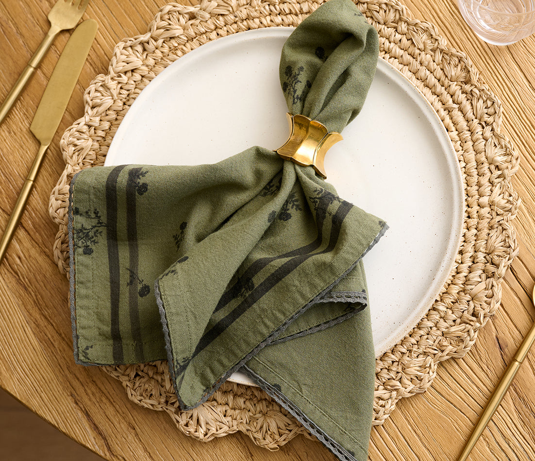 An olive green linen napkin with a dark floral pattern and gray border, folded and held in a gold ring on a white plate over a woven placemat.