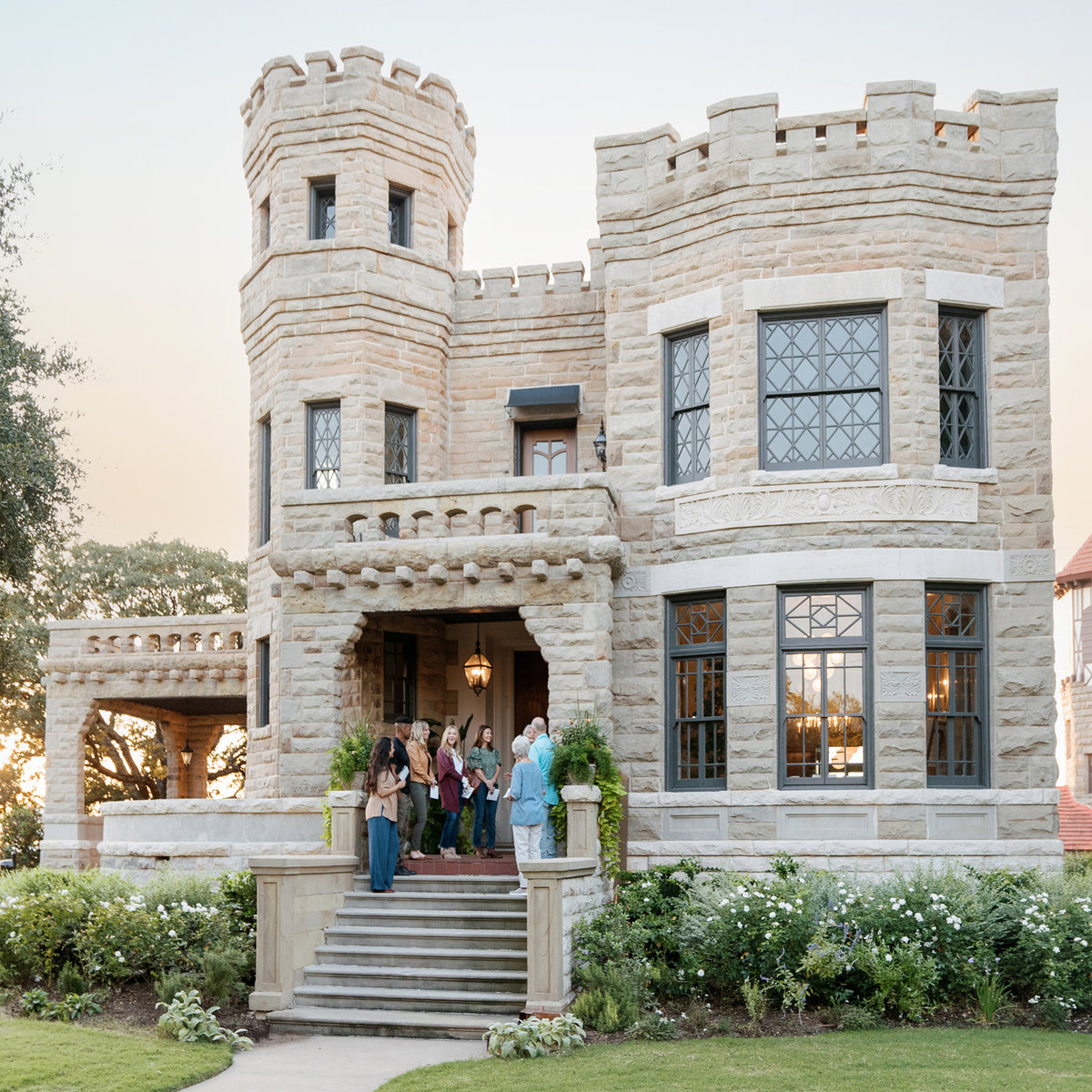 A large stone castle-style mansion with crenellated towers and dark-framed windows, shown at dusk with several people standing on the front porch steps and a few in a vehicle on the lawn.