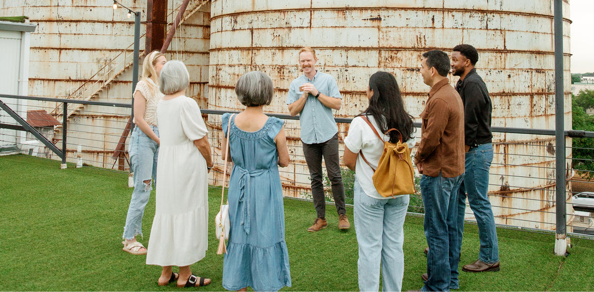 A group of seven people standing on a green rooftop in front of large, rusted metal grain silos, with a man in a light blue shirt speaking to the group.