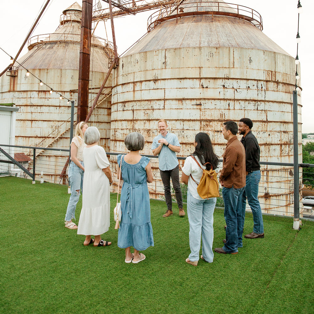 A group of seven people standing on a green rooftop in front of large, rusted metal grain silos, with a man in a light blue shirt speaking to the group.