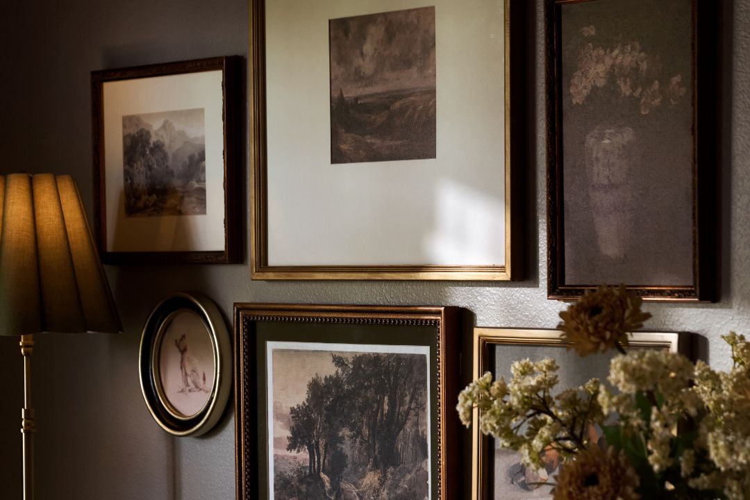 A gallery wall display featuring multiple framed art prints and paintings in dark wood and gold frames, styled with a lamp, flowers, and a textured wall.