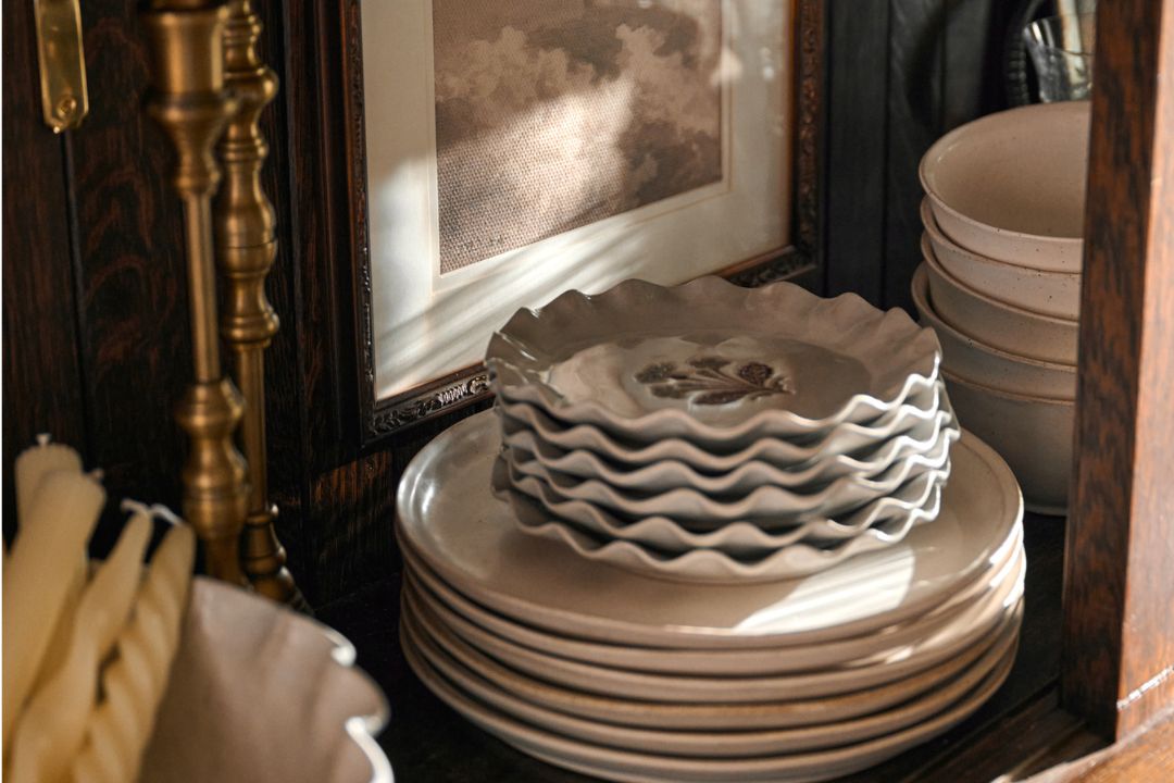 A stack of scalloped ceramic plates in a warm cream color, displayed on a dark wooden shelf next to a brass candlestick and framed artwork.