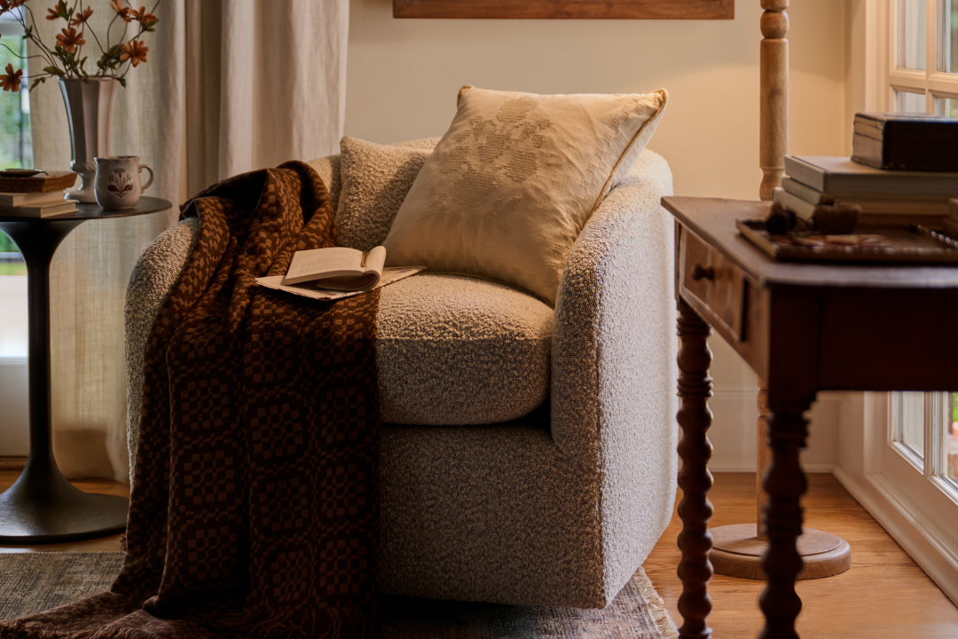 A cozy upholstered armchair in a warm beige bouclé fabric, styled with a cream pillow, a dark brown patterned throw blanket, and an open book on a wooden table in a sunlit room.