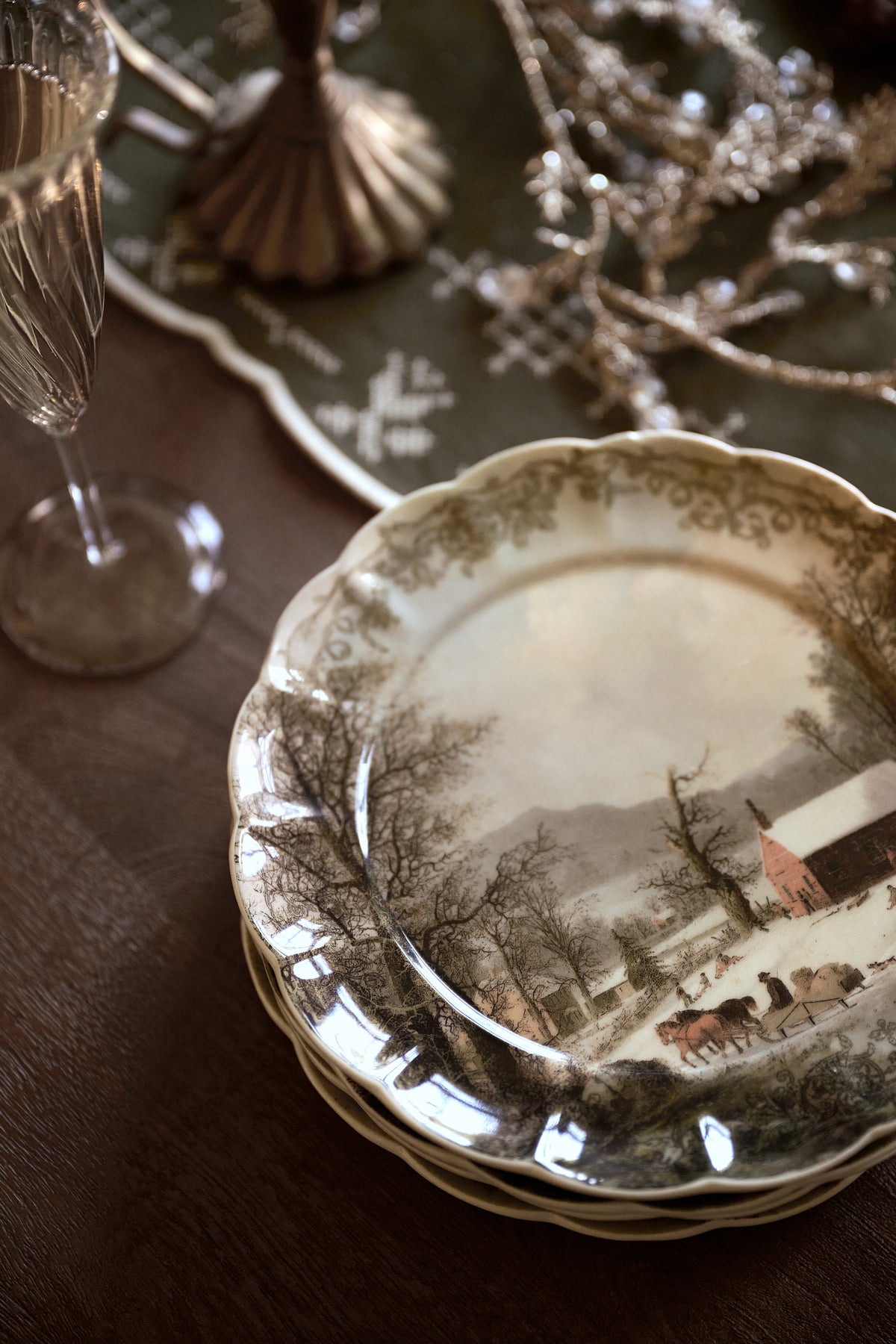 A stack of vintage ceramic plates with a delicate floral border and a winter landscape print, styled on a dark wooden table with a glass and silver holiday decor in the background.