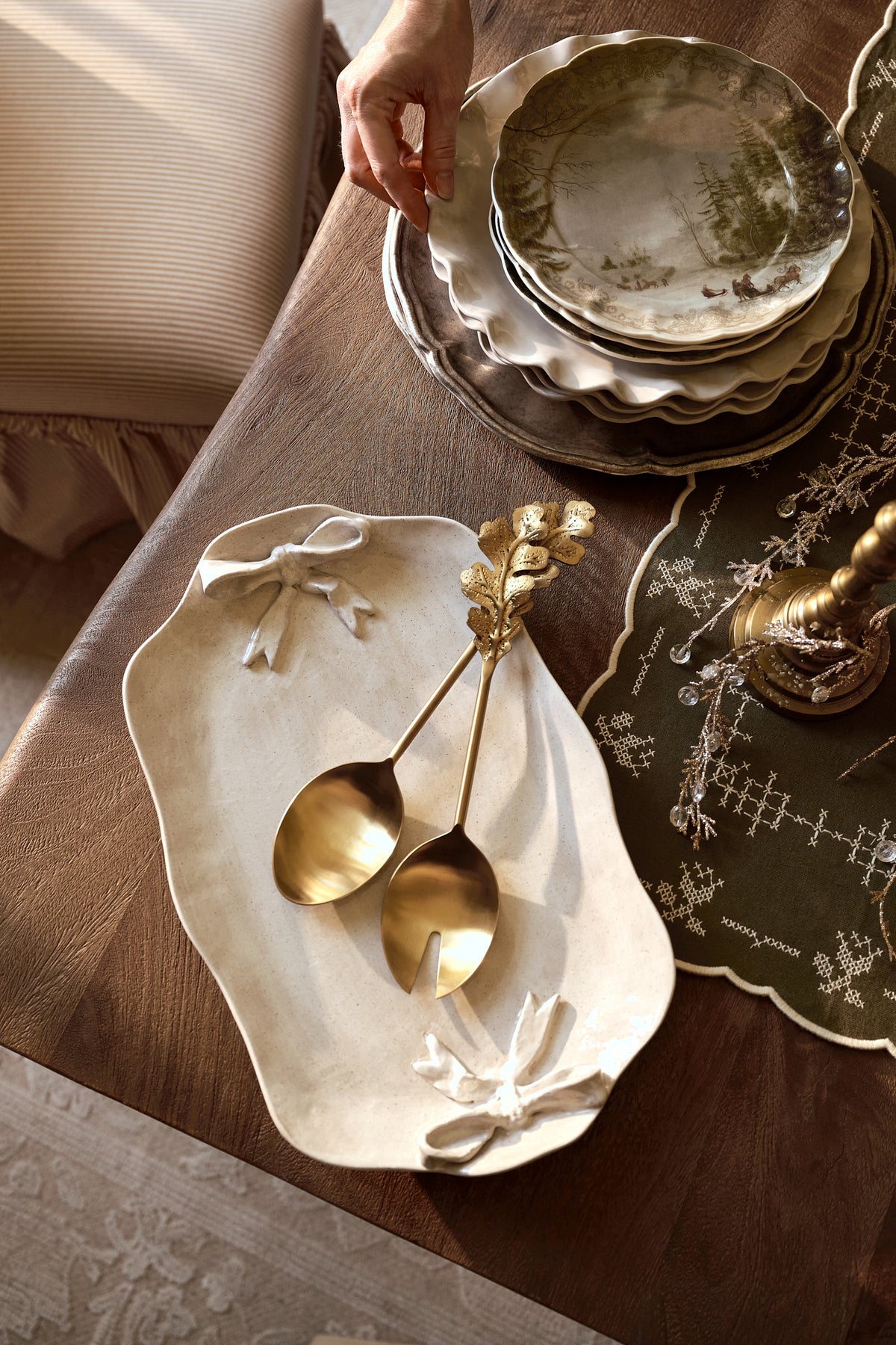A styled tabletop scene with a hand placing a ceramic plate on a stack of matching scalloped plates. In the foreground, a cream-colored sculptural serving platter holds two gold-toned spoons with floral handles, set on a dark wood table with a patterned green placemat.