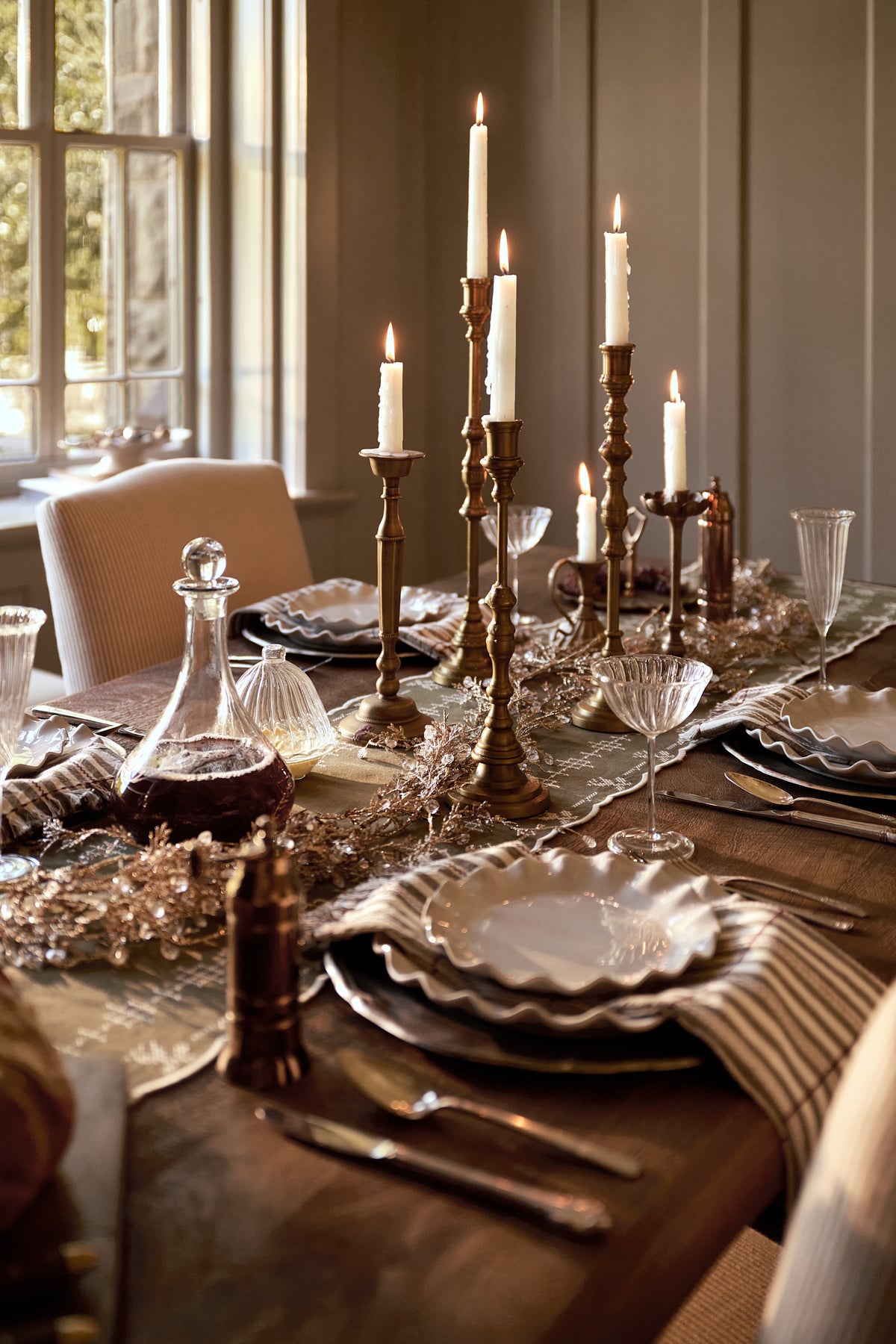A styled dining table set for a meal in a warm, elegant room. The table features layered white scalloped plates, silver flatware, a striped napkin, a clear glass decanter, and multiple brass and black candlesticks with lit white taper candles.