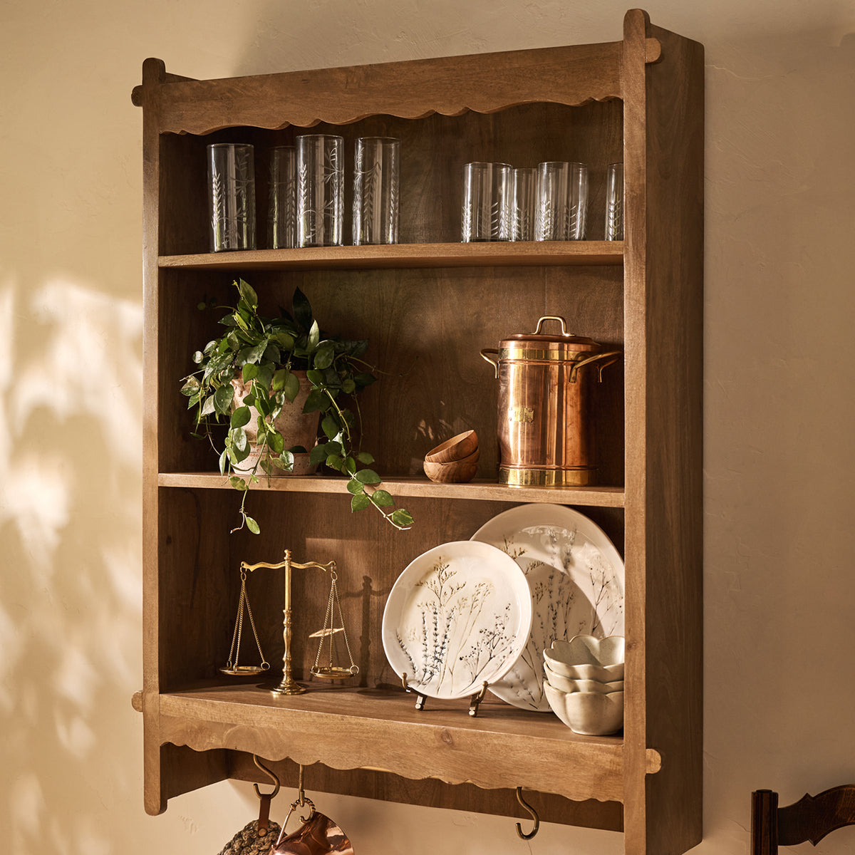 A rustic three-tier wooden wall-mounted shelf cabinet with scalloped edges, styled with glassware, a copper bucket, a potted plant, plates, bowls, and small decorative items against a beige wall.