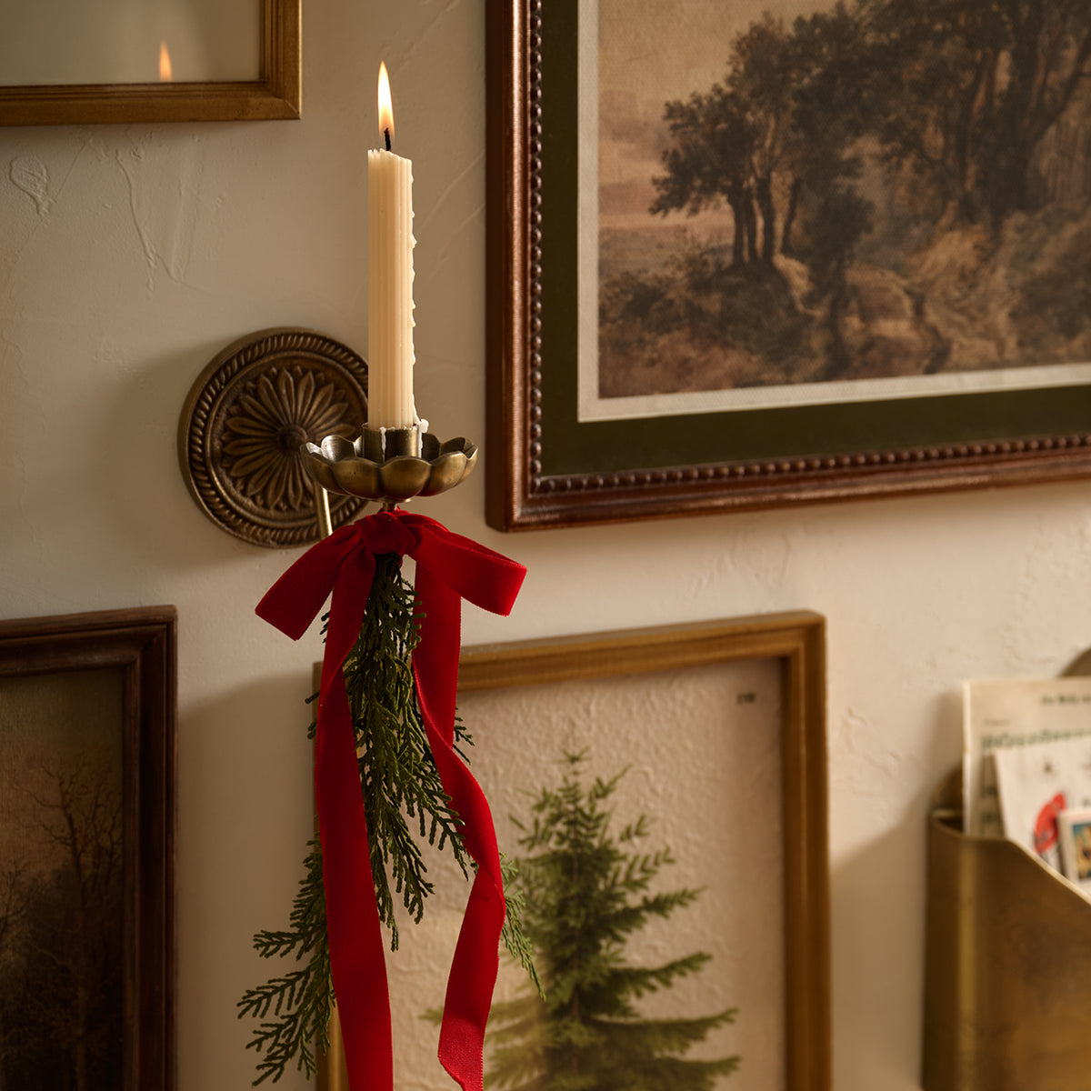 A lit white candle in a brass wall sconce holder, decorated with a red ribbon and green pine branches, mounted on a textured beige wall with framed artwork in the background.