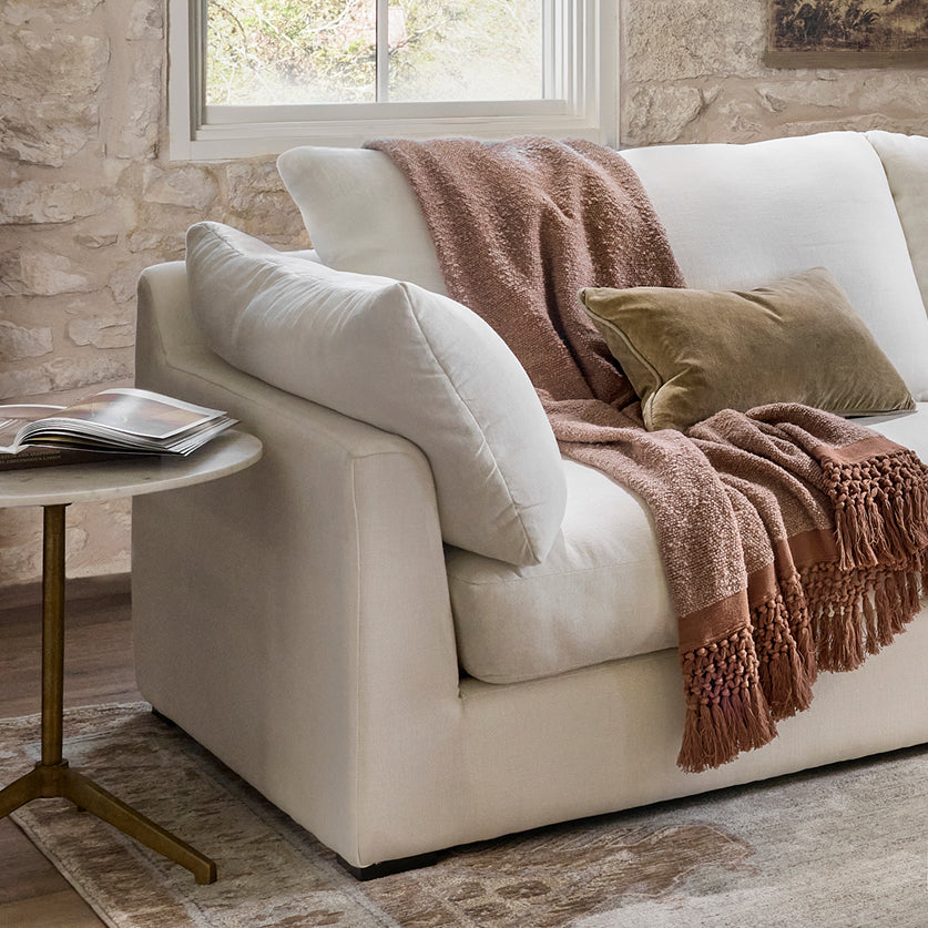 A cream-colored upholstered armchair in a rustic living room setting, styled with a brown textured throw blanket, a matching brown pillow, and a small side table.