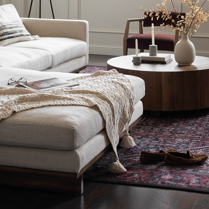 A styled living room scene featuring a cream upholstered ottoman with a chunky knit throw blanket draped over it, alongside a round wooden coffee table, patterned rug, and neutral-toned furniture.