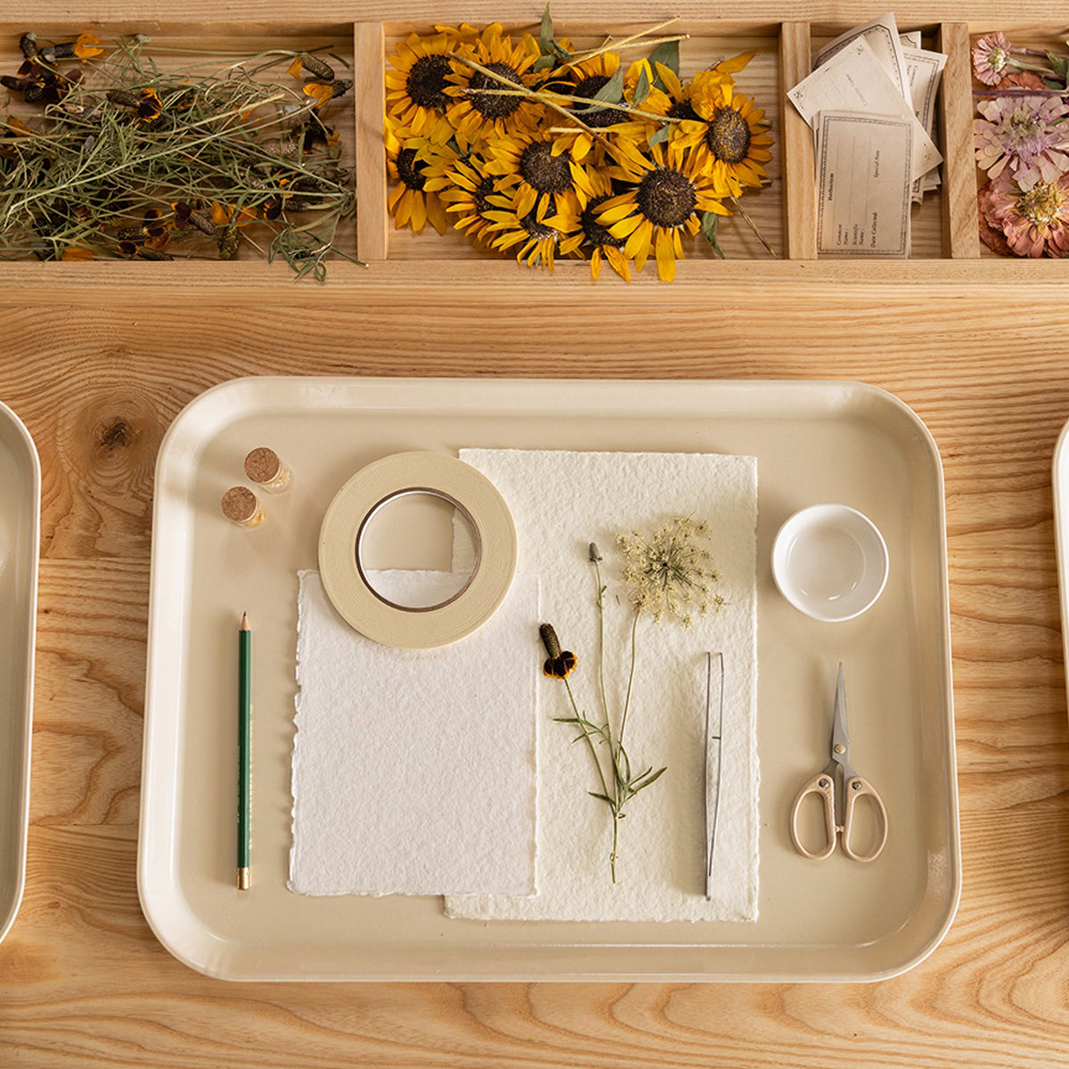A styled flat-lay of a beige tray containing art and craft supplies, including textured paper, tape, scissors, tweezers, and a small cup, set on a wooden table with dried flowers and sunflowers in the background.