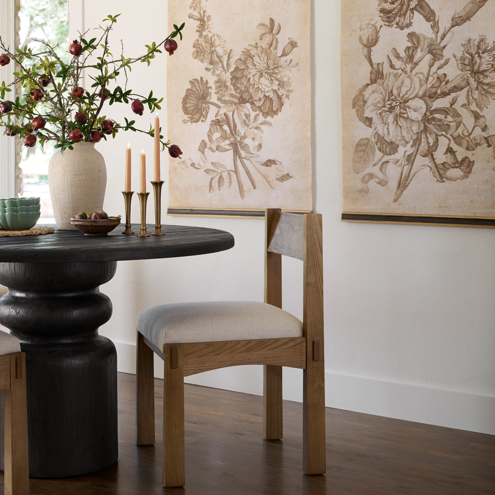Dining room with sepia-colored tapestries hung on the wall.  There is a rustic black wooden dining table with glass bowls, brass candlestick and a cream vase full of red fruit stems. There is a light wooden dining chair with cream upholstered seat pushed under the table.