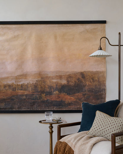 A large wall-mounted landscape tapestry with a black rod, hanging on a textured off-white wall in a styled room setting with a side table, lamp, and armchair.
