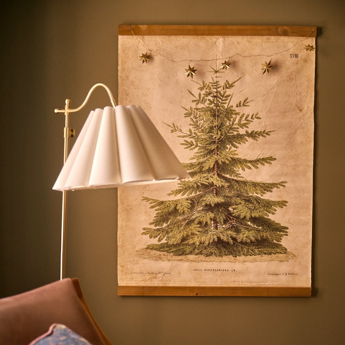 A holiday themed evergreen tapestry hung on a green wall with a scalloped lamp in front.