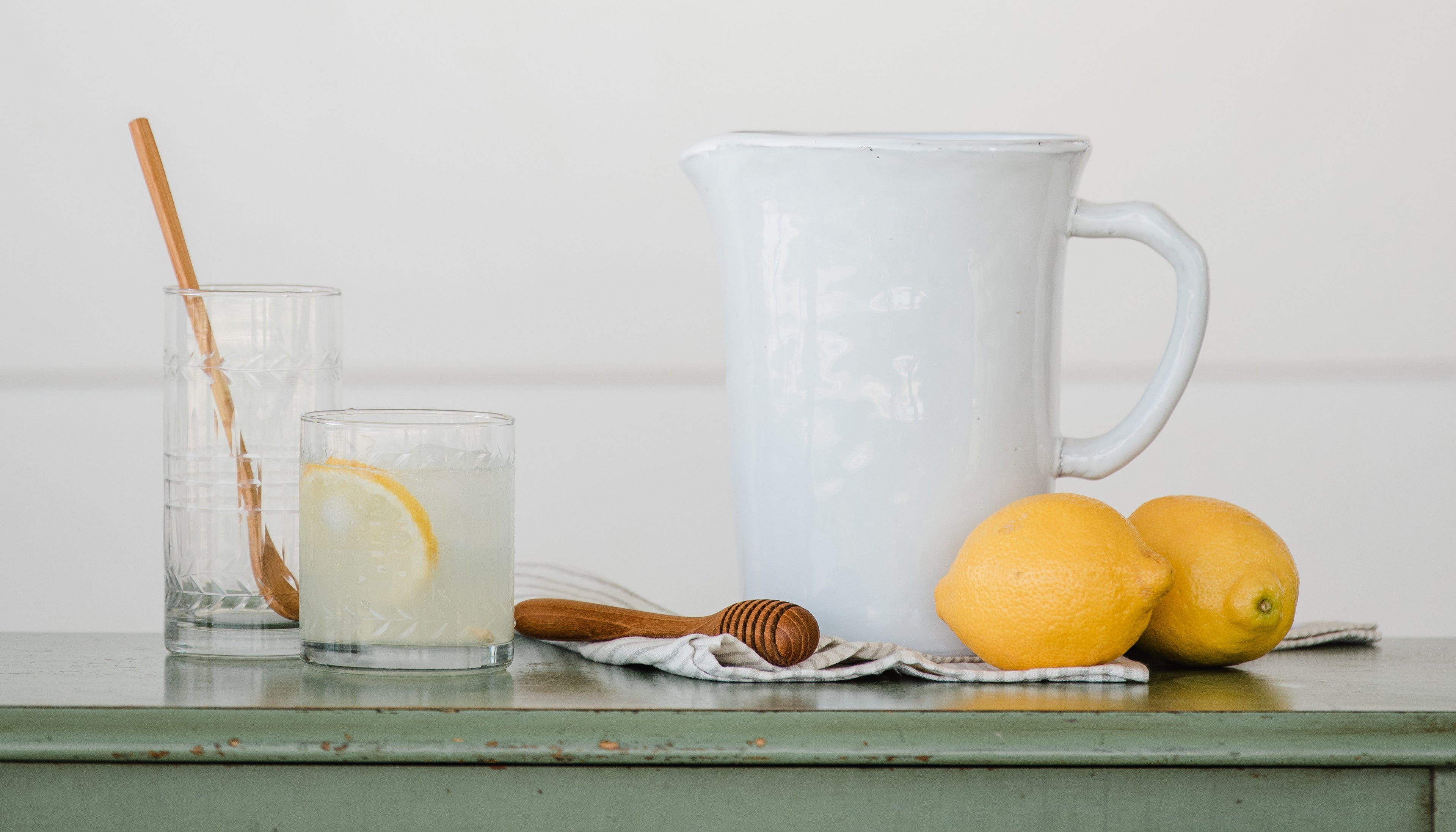 A white pitcher, kitchen tools, and a glass of lemonade sit on a green tabletop. 