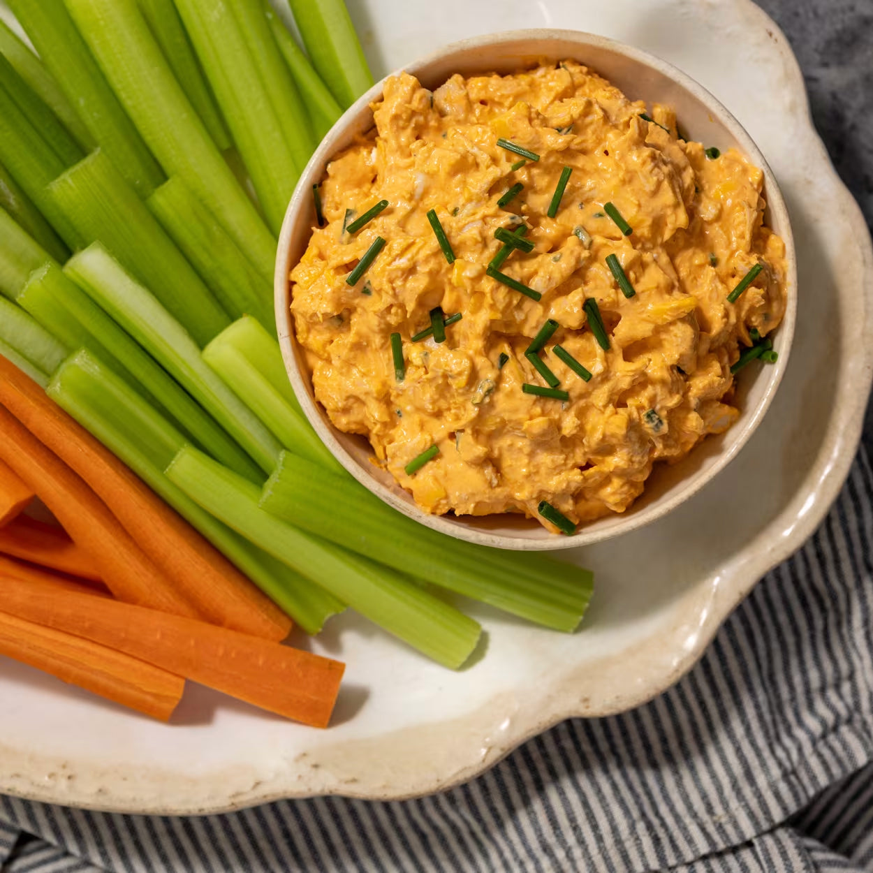 A bowl of chicken buffalo dip sits next to sliced carrots and celery.