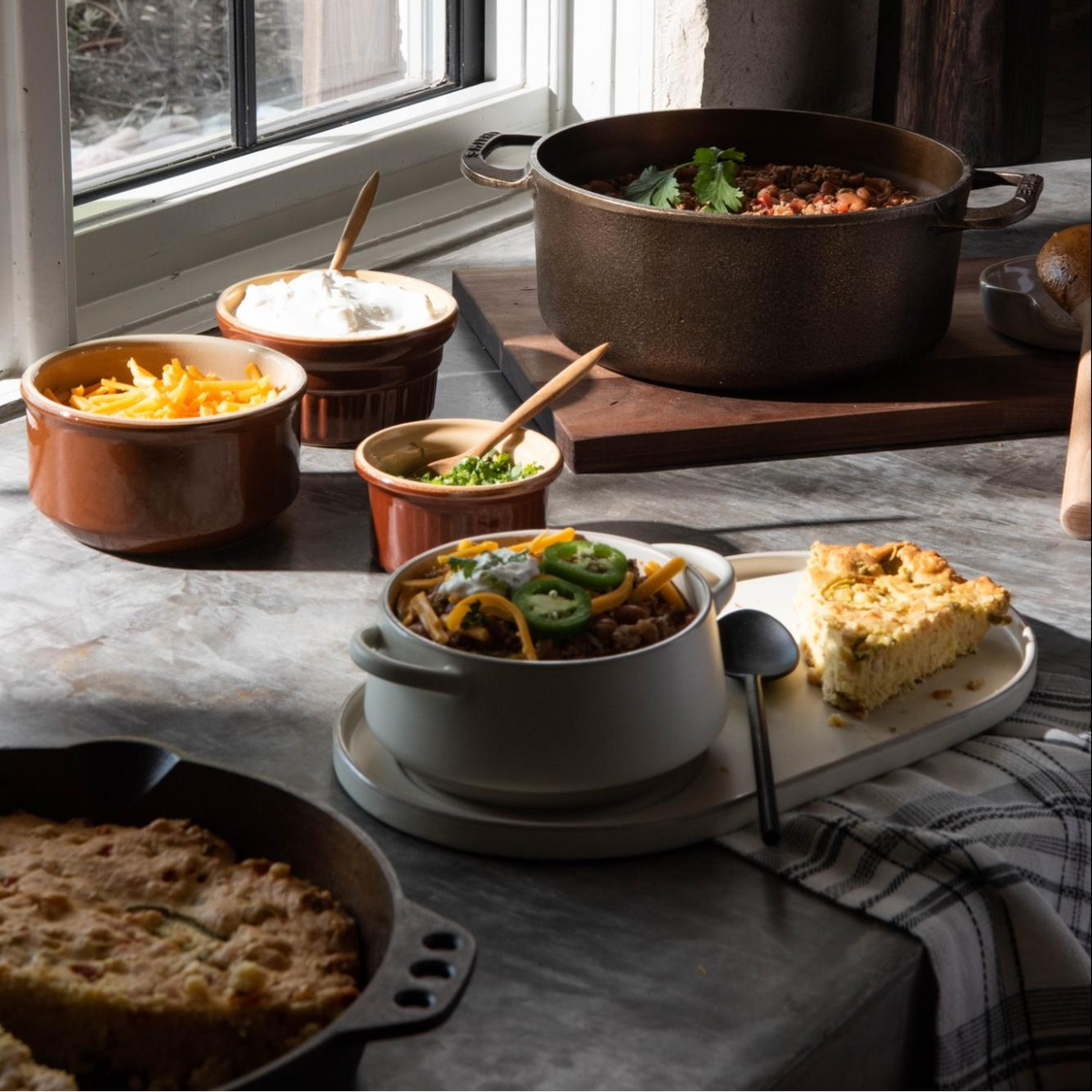 A rustic kitchen scene with a large dark cast-iron pot of chili, several brown ceramic bowls of cheese and sour cream, a white bowl of chili with jalapeño slices, baked cornbread, and a slice of casserole on a gray stone countertop near a window.