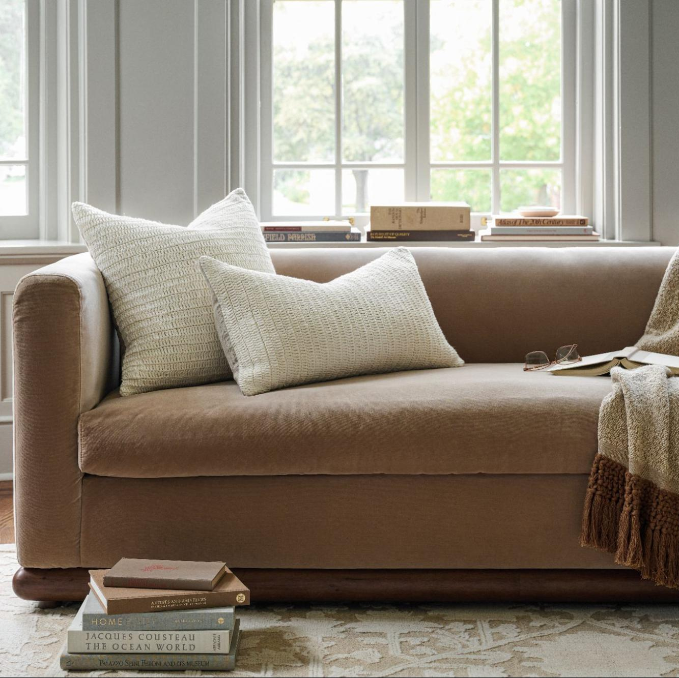 A brown upholstered loveseat sofa in a bright living room, styled with two cream textured throw pillows, a folded knit blanket, and a stack of books on the floor and windowsill.