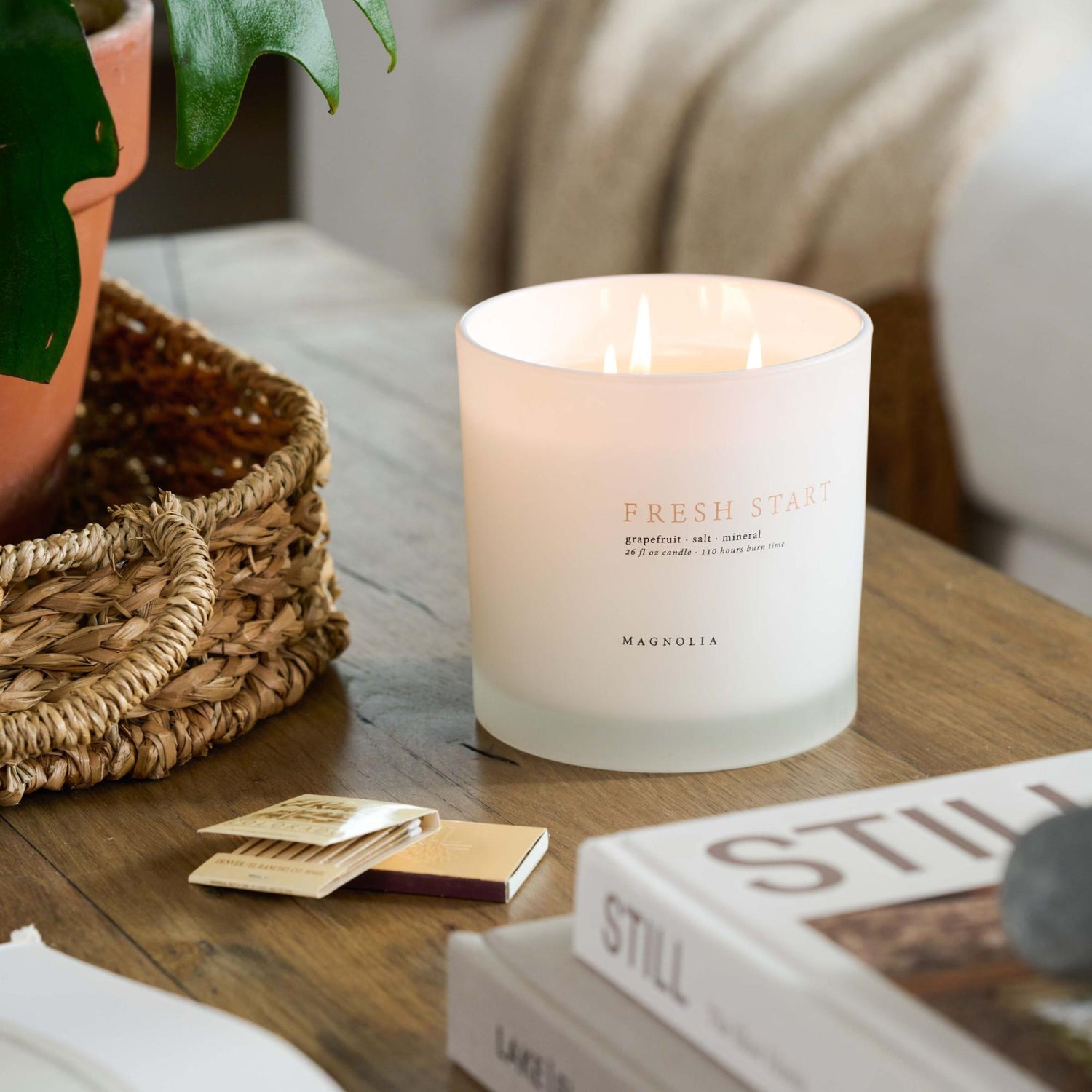 A flickering candle sits on a coffee table next to matches, books, and a basket.