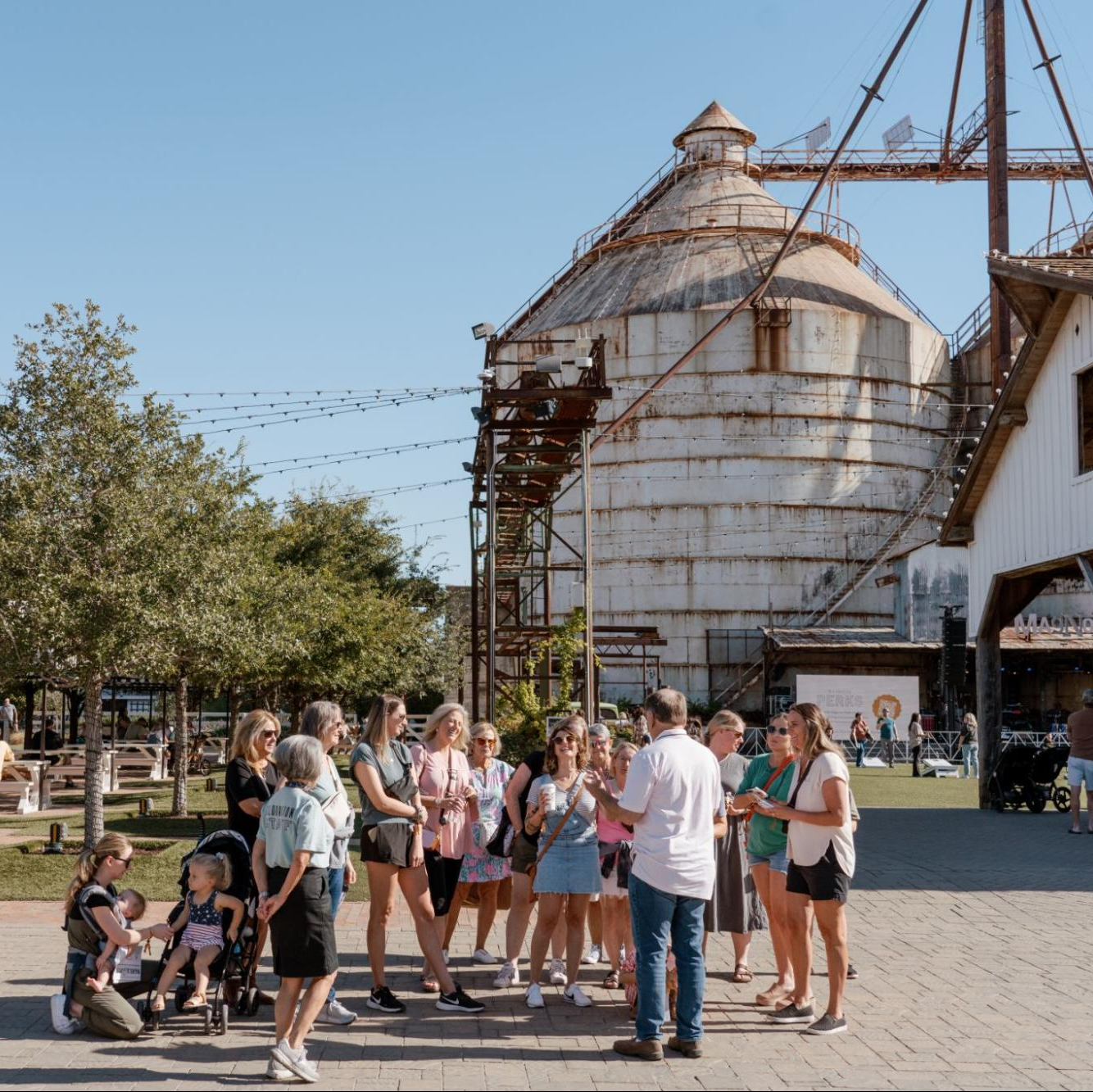 A group of people standing and talking outdoors in front of a large white cylindrical silo with rusted metal supports, under a clear blue sky. Trees, a building, and a paved walkway are visible.