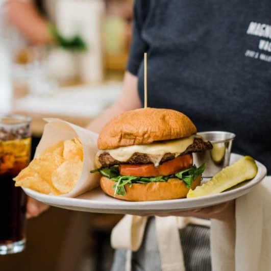 A close-up of a burger served on a white plate, held by a person wearing a dark gray t-shirt. The burger is topped with melted cheese, fresh green lettuce, and a tomato slice, with a wooden toothpick holding it together.
