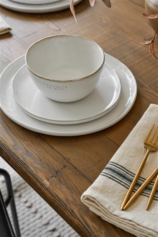 Two plates and a bowl from Magnolia sit on a wooden table next to a striped napkin and gold flatware.