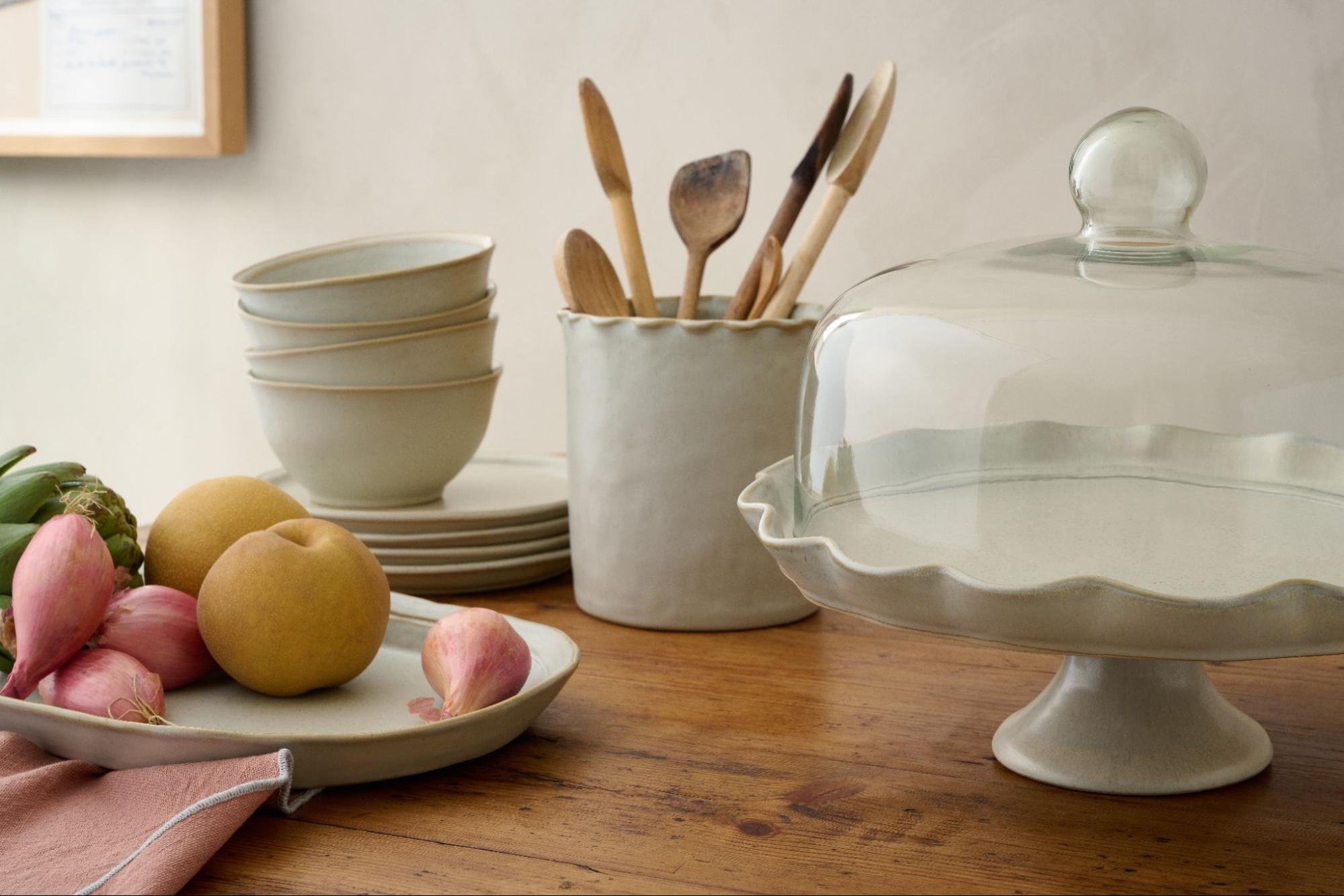 A stack of Magnolia’s French Grey Plates and Cereal Bowls sit on a wooden table—surrounded by a utensil crock, serving tray, and ruffle cake stand.