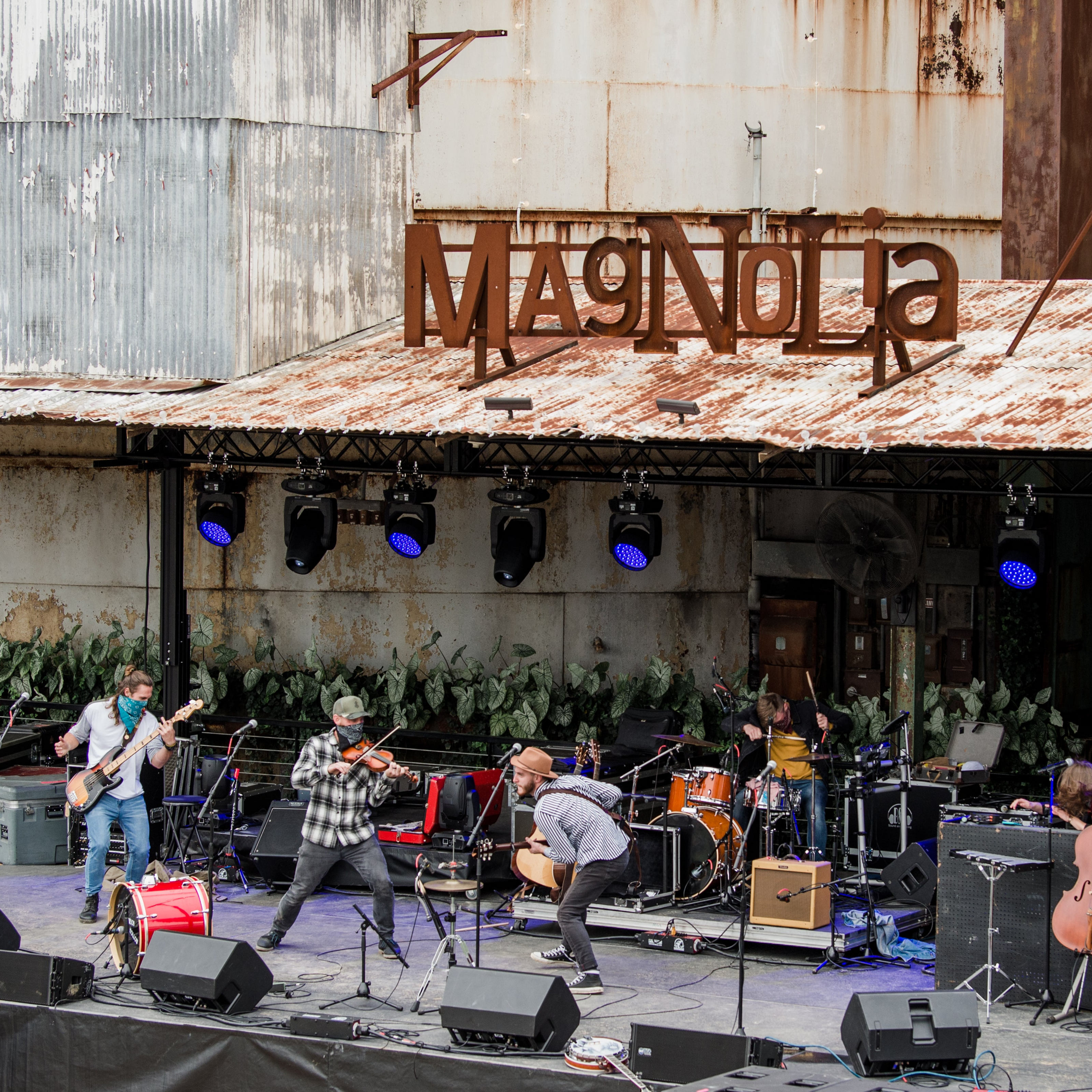 A live band performs on an outdoor stage in front of a rustic metal building with a large rusted 'Magnaolia' sign overhead. Musicians play instruments including guitars, a violin, drums, and a guitar, with stage lights and speakers visible.