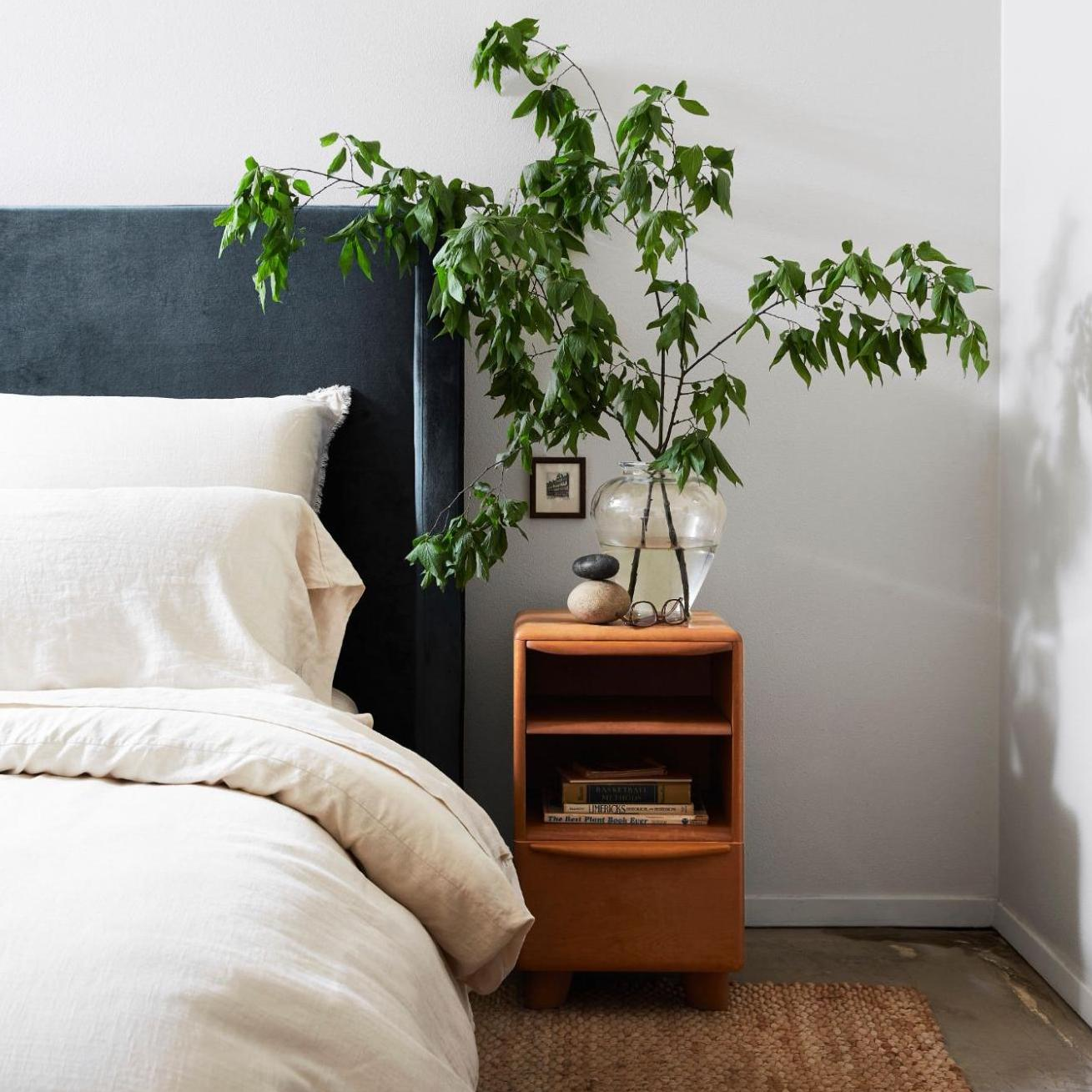 A tall green potted plant in a clear glass vase displayed on a wooden nightstand next to a bed with cream bedding and a dark gray headboard.