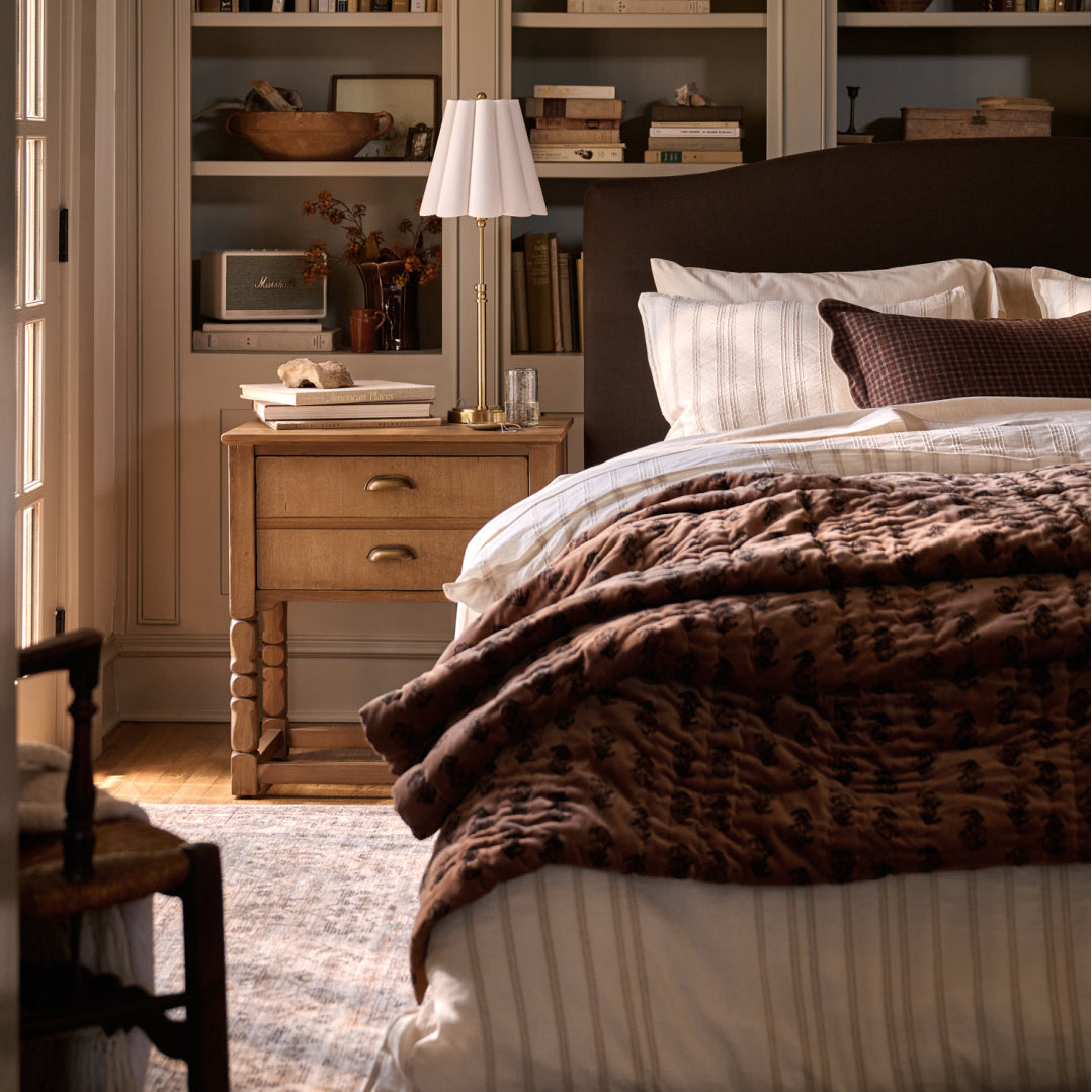 A styled bedroom scene featuring a bed dressed in white striped bedding and a textured dark brown patterned quilt, with a matching headboard, a wooden nightstand, a table lamp, and built-in shelving in the background.