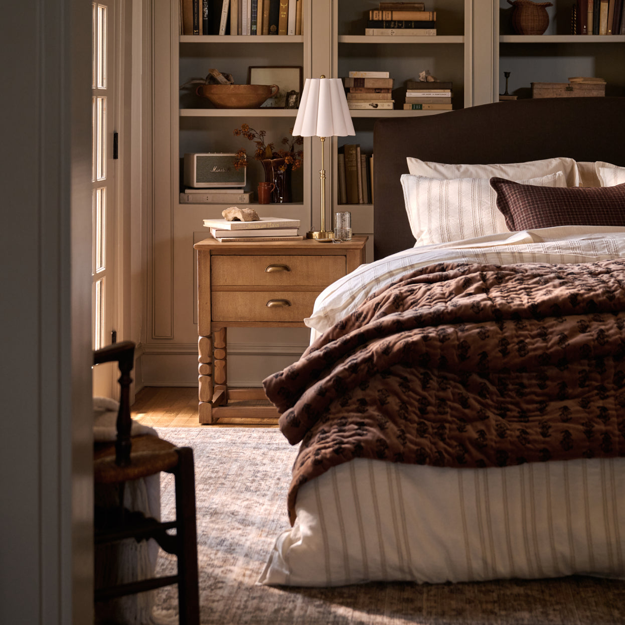 A cozy bedroom scene featuring a bed dressed in white bedding and a textured brown throw blanket, with a wooden nightstand and lamp beside it and built-in bookshelves in the background.