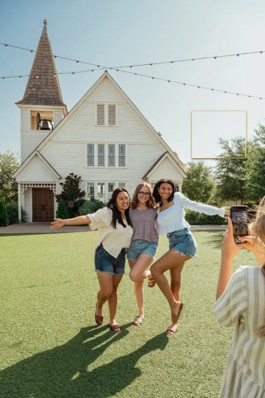 Three young women are jumping and posing for a photo outdoors on a green lawn in front of a white church building. A person on the right is holding a phone to take the picture.